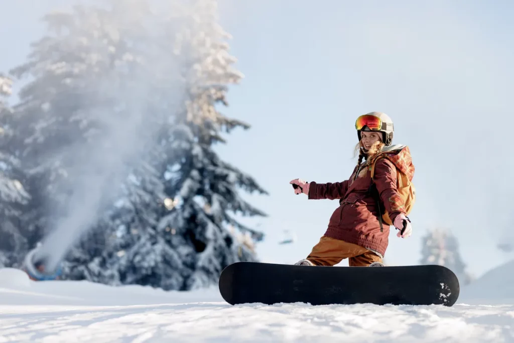 A person rides a snowboard down a snowy slope. Snow-covered trees are seen in the background. The sun shines brightly as the skier wears a helmet and protective gear.